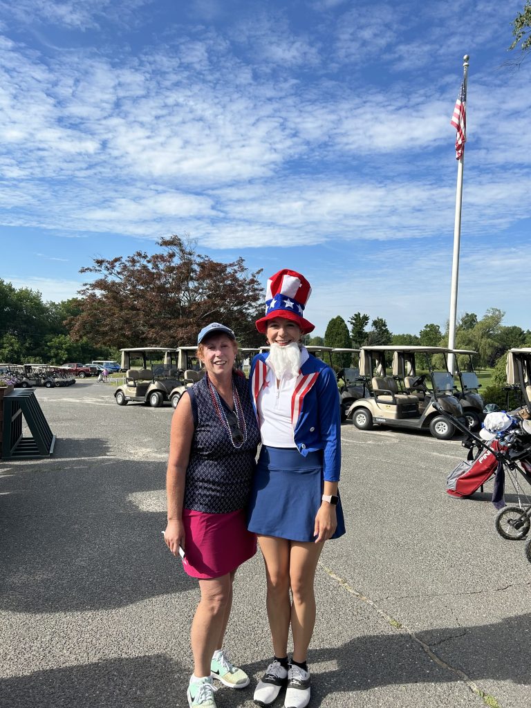 2 women smiling for the camera on a sunny day at the course parking lot. One in golf attire and the other in an Uncle Sam costume.