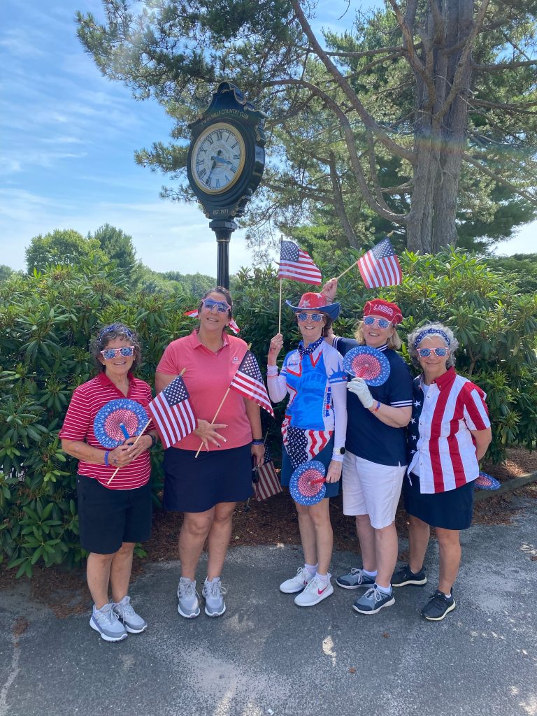 5 women celebrating the Fourth of July at the course, posing in front of the clock post. Everyone is wearing red and blue with sunglasses and holding American flags.