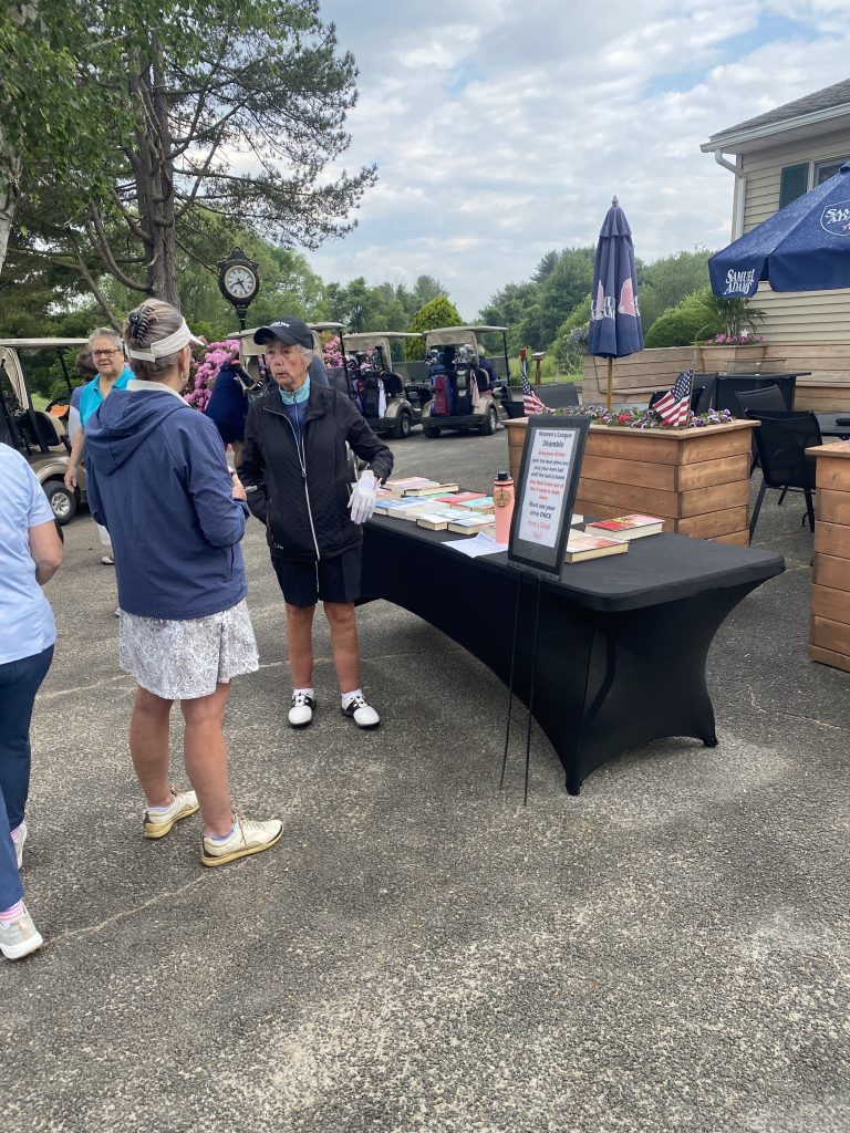 Women golfers line up in front of a booth right outside the clubhouse on a sunny day.